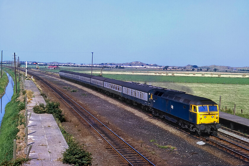 47487, 09.10 Holyhead-Manchester Victoria (1J16), Talacre 
 A photograph that shows the real and tangible weakness of Fujichrome slide film! Whilst I spent a long time in Photoshop working on the digital scanned image it still has a strange colour hue that I could not quite eradicate. The view is taken from an overbridge that leads down to the holiday village of Talacre and shows 47487 heading east with the 09.10 Holyhead to Manchester Victoria service. How warming to see a seven-coach locomotive hauled train working this 1J16 service that today would be operated by some two or three-car DMU; how things have changed! Notice the concrete platforms of the closed Talacre station that was shut in 1966 along with a number of others along this route. The platforms are still in place today but no holiday trains carrying passengers to the nearby vast Talacre Beach Resort holiday park will call again anytime soon! 
 Keywords: 47487 09.10 Holyhead-Manchester Victoria 1J16 Talacre