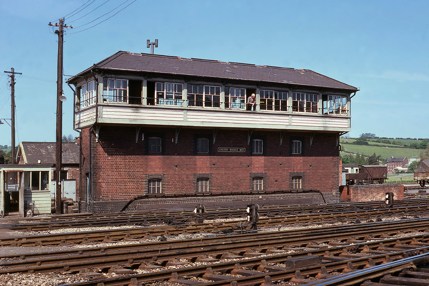 Exeter Middle Signal Box (GW, 1914) 
 With the signalman taking a breather and basking in some springtime sunshine, the grand Exeter Middle signal box is seen. It is a GWR Type 31 structure dating from 1914 and contained a 95 lever frame. Notice the diminutive crossing keeper's hut to the left of the box. The crossing keeper spent his day managing Red Cow level crossing that could cause huge congestion, particularly on a busy summer Saturday. Behind the box to the far right are two vintage wagons. There is a 12 ton twin-axle fruit van next to a coach and then a clay hood that would be making its way from the china clay quarries in Cornwall. 
 Keywords: Exeter Middle Signal Box