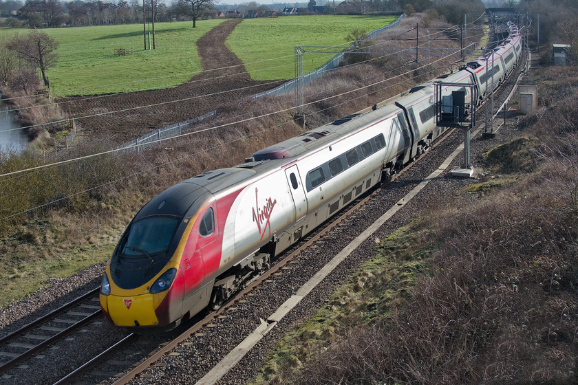 390129, VT 11.00 London Euston-Manchester Piccadilly (1H20, RT), A45 Weedon bypass bridge 
 The early spring sunshine catches the side of 390129 'City of Stoke-on-Trent' sweeps around of series of reverse curves at Weedon in Northamptonshire. The Pendolino is forming the 11.00 Euston to Manchester Piccadilly and its tilting mechanism will be in full action on this section of line to avoid the cappuccinos and skinny flat whites being spilt! Notice the strange area of exposed soil in the field behind the train. This was where contractors who were involved in the building of the bridge and bypass had one of their 'camps' and its associated access road. This will soon become part of the field again with no reference to them ever being there. 
 Keywords: 390129 11.00 London Euston-Manchester Piccadilly 1H20 A45 Weedon bypass bridge