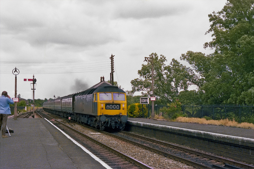 47456, unidentified down working, Bedwyn station 
 47456 opens up as at speeds through Bedwyn station with an unidentified down express for the West Country. The roof of the signal box can be seen above the locomotive. The 47 was a Midland interloper being a Crewe Diesel engine. Also, to the left Graham Vincent is seen taking his photograph behind his Velbon tripod that had his 6mm cin camera mounted on top. 
 Keywords: 47456 down working Bedwyn station