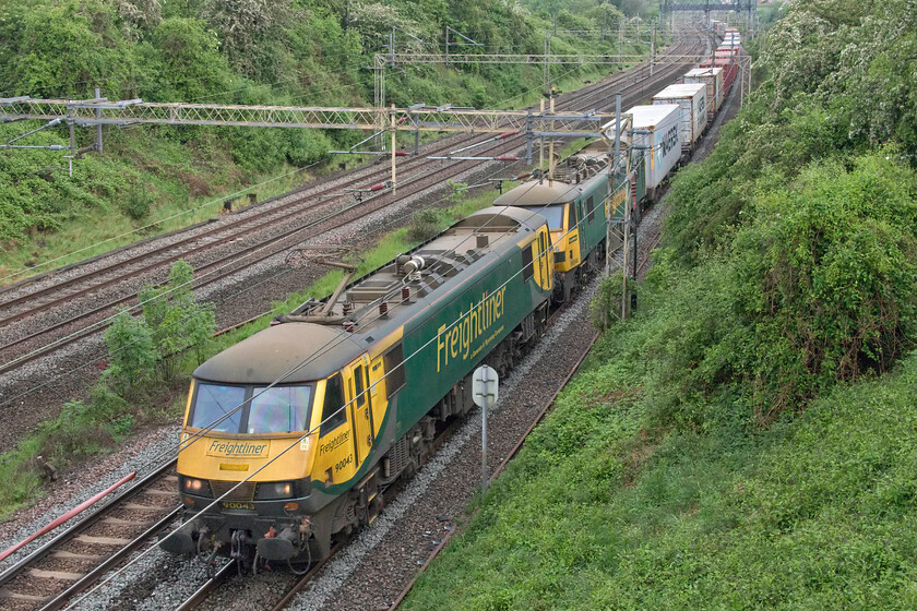 90043 & 90046, 04.10 Crewe Basford Hall-Felixstowe North (4L89, 4E), Victoria bridge 
 On a dull spring day with predominantly green surroundings, a pair of Freightliner liveried Class 90s do not stand out particularly well! 90043 and 90046 make light work of the 4L89 04.10 Basford Hall to Felixstowe service as it passes Victoria bridge just south of Roade. It is noticeable how much faster the electric hauled services are rather than those operated by a Class 66 with them taking significantly less time to reach this location from Northampton some seven miles away. 
 Keywords: 90043 90046 04.10 Crewe Basford Hall-Felixstowe North 4L89 Victoria bridge Freightliner