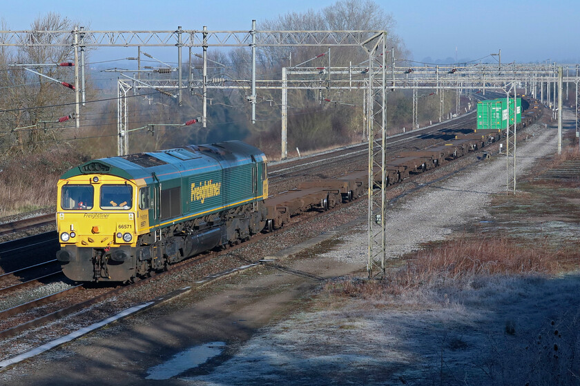 66571, 03.14 Garston-Felixstowe North (4L97, 2E), Gordon's Lodge 
 In perfect lighting, the 4L97 03.14 Garston to Felixstowe North Freightliner passes Gordon's Lodge between Northampton and Milton Keynes led by 66571. Notice the heavily frosted ground in the foreground as a result of the very cold and clear night that had just passed. Also notice what appears to be the driver's hands resting on the top of the control console. However, close examination of the RAW file reveals that they are, in fact, a pair of orange 'hi-viz' gloves! 
 Keywords: 66571 03.14 Garston-Felixstowe North (4L97, 2E), Gordon's Lodge Freightliner