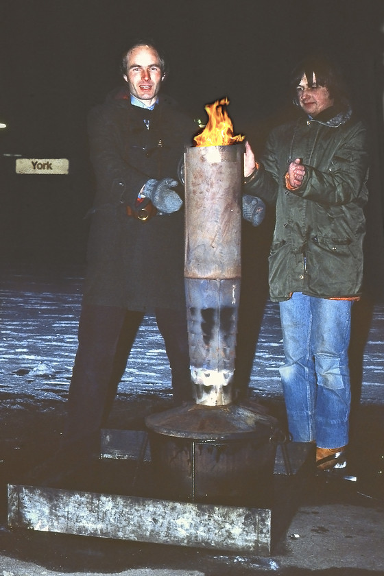 Graham & myself warming ourselves, York station 
 Could you even begin to imagine equipment like this being left on a railway station today? This incredible piece of infrastructure was composed of a tin bath containing paraffin or some sort of oil that was simply being burnt in this huge chimney type structure with flames emerging from the top. They were placed on the platform ends near to where the standpipes were located that were used to replenish the steam heat boilers of the locomotives with the aim of preventing the water pipes freezing during bitter winter nights such as this one. As can be seen from the lack of snow around them they appeared to be doing an effective job! Graham and yours truly took full advantage of their warmth gathering around them at this unearthly hour on York station. Whilst Graham has gone for the old dependable Duffle coat I have opted for the period green Parka jacket complete with faux fur lining to the collar and hood! Happy days! 
 Keywords: Graham York station