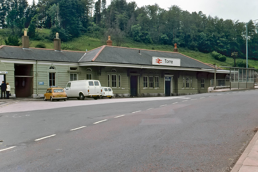 Frontage, Torre station 
 The frontage at Torre station is seen on a quiet Sunday afternoon, with just three cars in the car park, one of which is our own orange Mini. The timber-framed building, clad with horizontal boarding, was opened by the GWR in 1848 when just one platform served a single broad gauge track. When standard gauge replaced the broad gauge in 1892, a second platform was added to serve the new up line. The station is Grade II listed and looks very similar to this today, but the road in the foreground is a little busier and much of the station is now split up into small business units. 
 Keywords: Frontage Torre station