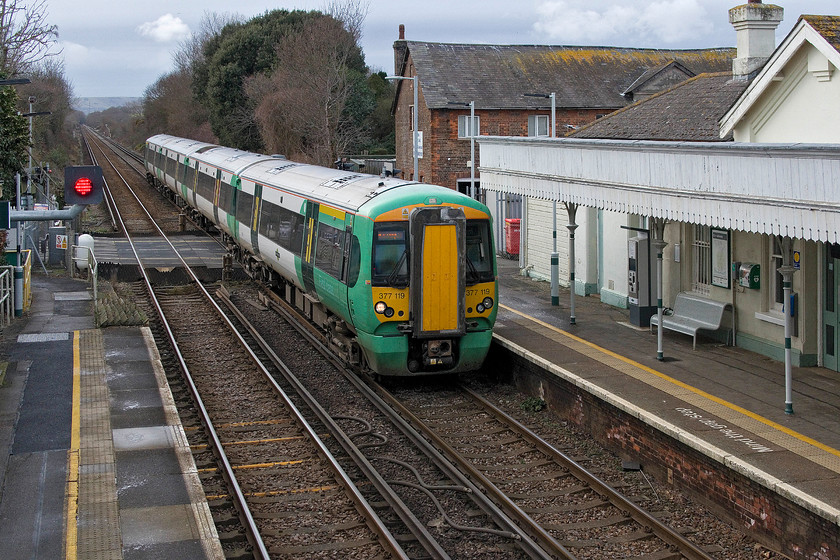 377119, SN 13.31 Brighton-Hastings (1G36, RT), Pevensey & Westham station 
 377119 passes through Pevensey and Westham station with the 13.31 Brighton to Hastings service. Notice the relay room on the down platform surrounded by palisade fencing, this was where the signal box was located. Also notice the white RADAR/LiDAR transmitter/receiver by the level crossing. This is part of the object detection system that is one control aspect of the crossing. It is this system that gave so much trouble when it was installed on the Thetford line particularly at Brandon level crossing. 
 Keywords: 377119 13.31 Brighton-Hastings 1G36 Pevensey & Westham station