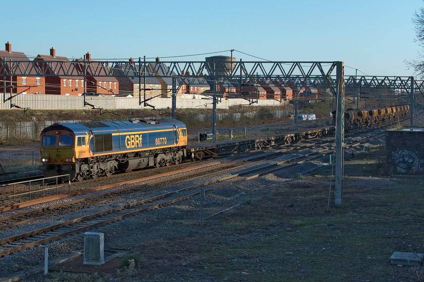 66770, 13.15 Milton Keynes Central-Bescot (6G51, 8E), site of Roade station 
 After an overnight possession of the slow lines south of Hanslope Junction an infrastructure working heads back to its base at Bescot as the 6G51 13.15 from Milton Keynes. Behind the flats are a number of HQA (E) eighty-eight tonne auto discharge ballast wagons that were empty, indicating that there will be some fresh ballast at the worksite. The train is seen ambling past the site of the former station at Roade. 
 Keywords: 66770 13.15 Milton Keynes Central-Bescot 6G51 site of Roade station GBRf