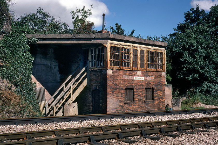 Stoneycombe (Sidings) signal box (GW, 1894) 
 I cannot fully remember how Graham and I got to this position opposite Stoneycombe Sidings signal box, to give it its proper and full name despite what the BR corporate image sign says. I think that it involved climbing some fences and a walk up the line, something that could and should never be undertaken today. Surprisingly, despite appearances, the box was a relatively early construction dating from 1894 and has an unusual flat reinforced concrete roof to protect it from falling stone during blasting. There was a huge hole in the ground just behind it where the stone had been extracted over the years with an even larger one behind where I am standing to take the photograph. Whilst the signal box could still be operated it rarely was being semi-permanently switched out. Amazingly, the box still stands with the widows bricked up and somewhat enveloped in greenery and can be seen, if one is quick, from passing trains. The frame still survives and is in use at the GWR Didcot Railway Museum now fitted to the former Frome North box now named Frome Mineral Junction. 
 Keywords: Stoneycombe Sidings signal box GWR Great Western Railway