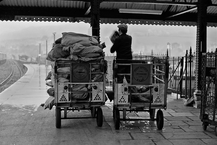 Sorting mail bags, Bath Spa station 
 A sight that was once so familiar that has totally disappeared from the railways, the conveyance of Post Office mail. Complete with his cloth cap an employee sorts bags of mail for onward movement either down under the station for further sorting or for transfer to a train. As seasoned spotters, we were always pleased to see an empty Post Office trolley on the platform end as it made an ideal place to sit, spread out one's kit and settle down to collect numbers. They were infinitely better than a BRUTE trolley seen to the right as these were too low down. However, I seem to recall that the Post Office trolleys had a vertical ridge running along the open side that ended up digging into the back of one's thighs after sitting on a trolley for too long! 
 Keywords: Sorting mail bags Bath Spa station The Piost Office