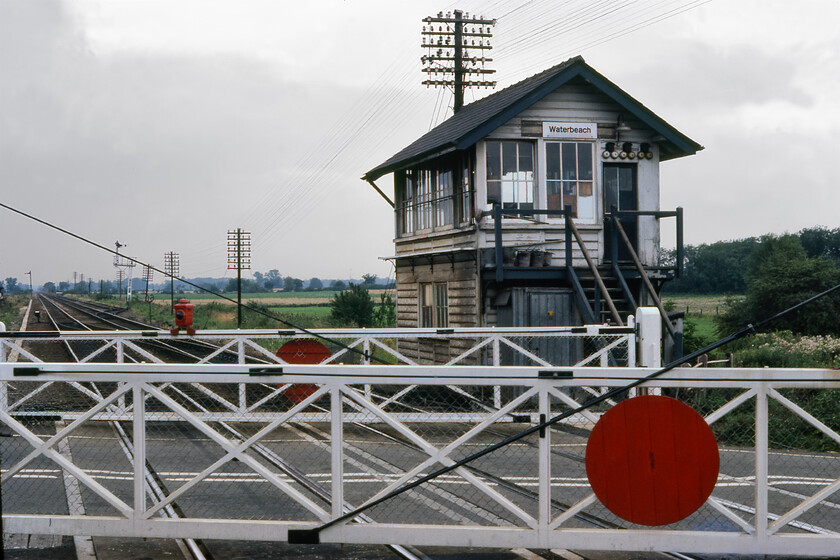 Waterbeach signal box (GE, 1882) 
 The fortunes of Waterbeach station have gone through many changes over the years. Today it is a relatively busy commuter station just over sixty miles from London with passenger numbers on the ascendancy after the COVID dip. The platforms are now extended and staggered being either side of the level crossing as well as the route now being electrified. This means that in the 1981 view of the former signal box the up platform is now located to the left beyond the level crossing. The 1882-built Great Eastern Type 2 looks a little tatty which is not surprising really as it was set to close just four years after this photograph was taken. Notice the six bells on the gable end above the door that would have created quite a crescendo when the gates were to be closed. The saga of Waterbeach station is not yet over as there are now controversial plans to relocate it further north in response to plans to develop a New Town of between eight and nine thousand homes on the former Waterbeach Barracks. 
 Keywords: Waterbeach signal box GE Great Eastern Railway