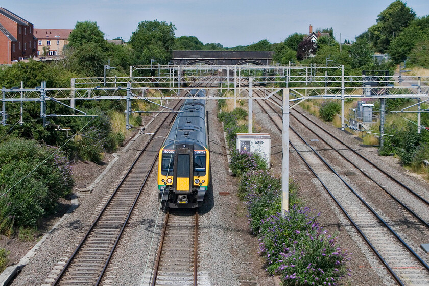 350265, LM 10.02 Crewe-London Euston, site of Roade station 
 Running 'fast' in the second part of its journey, 350265 passes Roade working London Midland's 10.02 Crewe to Euston service. Hard to believe that there was, until 1964, a four-platform station occupying the spot where the Desiro is passing. All platforms were linked by a footbridge approximately where the second electrification pylon is that linked across to a small station building where the greenery is on the far right. The station master's house still stands with its gable end and chimney to the top of the photograph. 
 Keywords: 350265 10.02 Crewe-London Euston site of Roade station