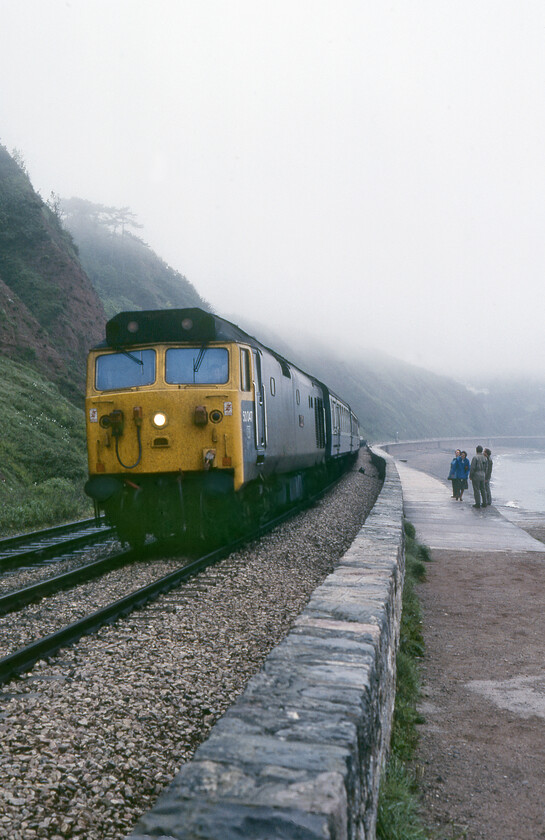 50047, 07.45 Kensington Olympia-Newton Abbot & St. Austell (1V34), Teignmouth 
 Watched by a group of 'normals' out for a promenade on the rather damp and foggy sea wall at Teignmouth 50047 'Swiftsure' makes a fine sight as it passes at such close quarters. 50047 was a refurbished member of the class identified by its centre-fitted headlight having been one of the earliest to be so treated entering back into service just under a year previously. It is leading the 1V34 07,45 Kensington Olympia to Newton Abbot and St. Austel Motorail service the flats of which can just be seen towards the rear of the train. The Newton Abbot portion of the flats will soon be detached on arrival in about ten minutes' time hopefully with the respective car owners doing the same! The remains of the train would then continue on into Cornwall. 
 Keywords: 50047 07.45 Kensington Olympia-Newton Abbot St. Austell 1V34 Teignmouth Swiftsure Motorail