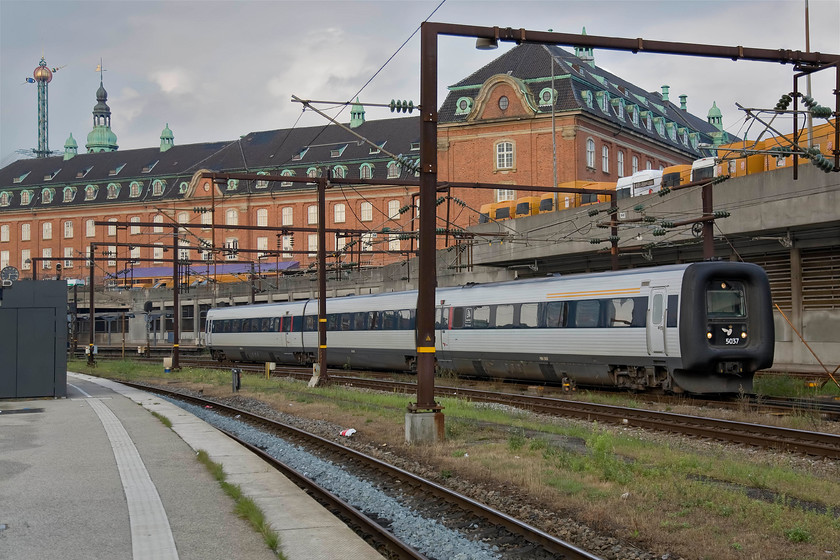 5037, unidentified working, Copenhagen Central station 
 I am not convinced about the design of the IC3 units as seen here at Copenhagen Central station. Their front end huge rubber diaphragms are designed to form an airtight seal when working in tandem with the whole of the driver's door and window folding away to provide a wide through gangway between sets. With the world-famous Tivoli Gardens Himmelskibet (En. Star Flyer) swing ride in the background, 5037 'Doctor Larsen' departs with an unidentified working. 
 Keywords: 5037 Copenhagen Central station Doctor Larsen