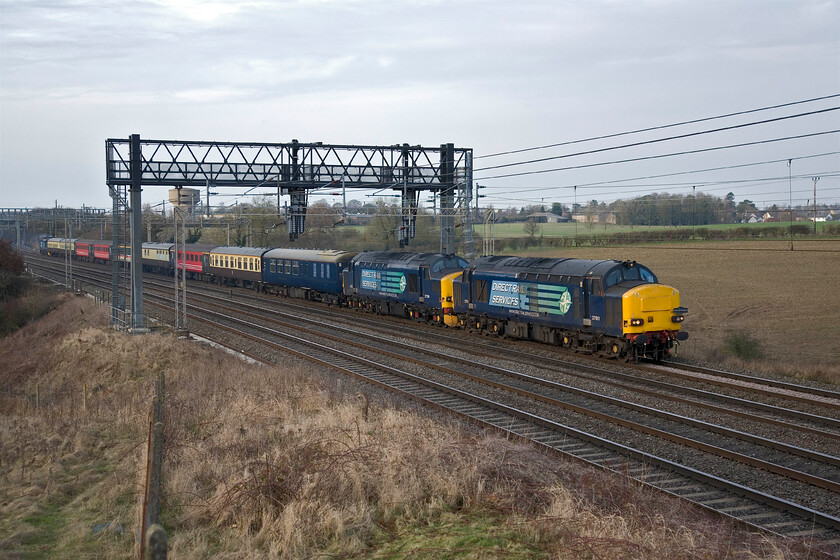 37611, 37259 (failed) & 37425, outward leg of The Enigmatic Logistition, 08.21 Crewe-Wolverton (1Z34, 2L), Roade Hill 
 At last! After an engine failure of 37259 (second locomotive) The Enigmatic Logistition charter has finally made some progress and is seen passing on the WCML just south of Roade in Northamptonshire. The excellent website Six Bells Junction documents the day's events well, see....https://www.sixbellsjunction.co.uk/ . The train is now led by 37611 with 37425 giving some power at the rear. Note the motley collection of stock with four different liveries in evidence. This photograph was published in Rail. 
 Keywords: 37611 37259 failed 37425 The Enigmatic Logistition 08.21 Crewe-Wolverton 1Z34 Roade Hill DRS Direct Rail Services