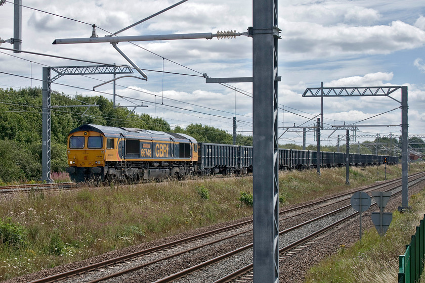 66748, 11.00 Acton Yard-Bardon Hill (6M46, 30E), Irchester SP927667 
 66748 'West Burton 50' trundles down from Sharnbrook summit on the down slow line at Irchester. The GBRf Class 66 is leading the 11.00 Acton to Bardon Hill empty stone train. Despite the up and down slow lines being re-laid last year, already there are extensive weeds growing between the rails at a point where the locomotive is in this photograph. Also in this image is the recently installed electrification hardware. Now hopelessly behind schedule, it has just been announced that this will not go live until, at the earliest, March 2021! 
 Keywords: 66748 11.00 Acton yard-Bardon Hill (6M46, 30E), Irchester SP927667 GBRf West Burton 50