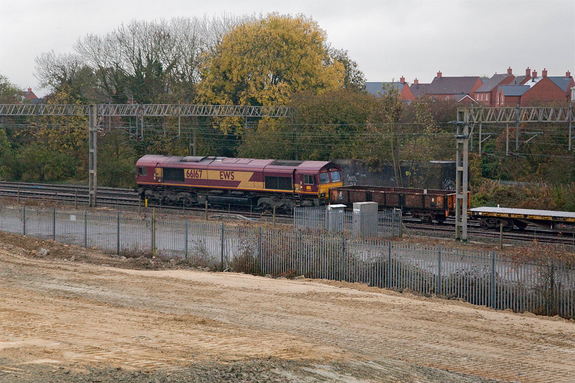 66167, 12.25 Wembley-Bescot, site of Roade station 
 66167 is seen on the rear of the 12.25 Wembley to Bescot engineers' train. The autumn colours are still evident in the trees behind that are still clinging on to their leaves despite it being early November. The houses in the background are just some of the new ones inflicted on the village of Roade with many more still to come on the cleared land in the foreground. 
 Keywords: 66167 12.25 Wembley-Bescot site of Roade station