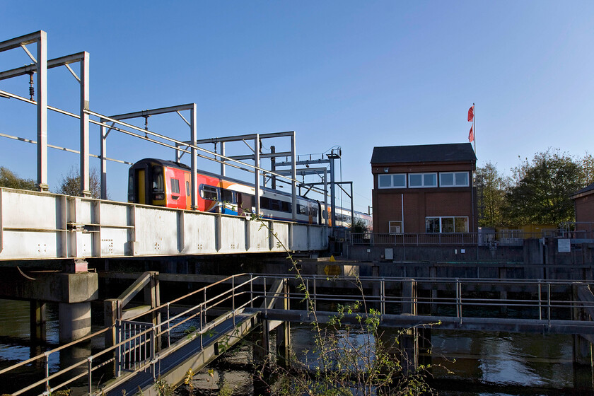 158854, EM 14.58 Norwich-Liverpool Lime Street (1R98), Trowse swing bridge 
 158854 heads away from its starting point working the 1R98 Norwich to Liverpool Limes Street service. It is crossing the single track Trowse swing bridge that spans the River Wensum that permits access to access to the historic port of Norwich that was established by an Act of Parliament in 1827. The signal box inspired structure on the other bank of the river controls the opening and closing of the swing bridge. 
 Keywords: 158854 14.58 Norwich-Liverpool Lime Street 1R98 Trowse swing bridge East Midlands Trains