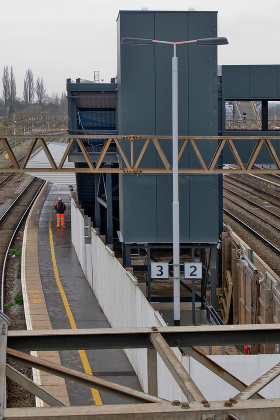 New footbridge, Northampton station 
 Under the beady eye of a worker on platform three, I take a photograph of the new station footbridge lift tower at Northampton station. During this work to fully rebuild the station, a temporary footbridge was built at the north end of the station and this can be seen through the first window frame of the new one complete with a passenger descending the steps. 
 Keywords: New footbridge Northampton station