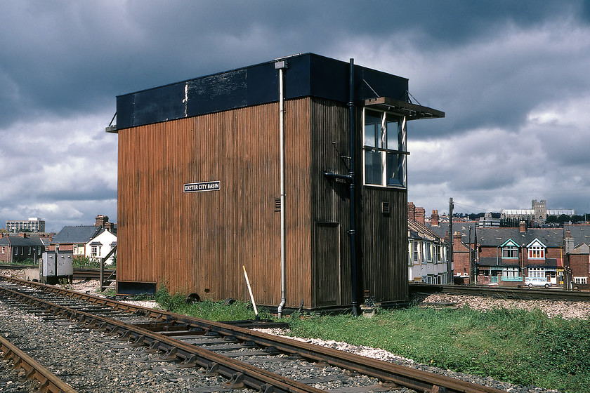 Exeter City Basin signal box (BR, 1962) 
 The rear of Exeter City Basin signal box is seen in some welcome sunshine This was one of the Western Region's 'plywood' boxes as they were often referred to being similar to others at Evesham and Onibury. This one was built in 1962 to British Railways and it is nice to see that even on the rear it retains a cast plate. This was as far as Graham and I dared get to the box as it was manned and we were the wrong side of the fence. The rusty tracks in the foreground led down to Marsh Barton but originally were at the start of the Great Western 'inland' route to Newton Abbot via Heathfield. With the continual pressure of the present sea-wall route today serious consideration is being given to the re0opeing of this route. Notice the magnificent Exeter Cathedral on the skyline. City basin box survived the initial resignalling of the Exter area in 1985 surviving on another year until November 1986 to permit railway access to Marsh Barton with the demolition gang moving in during February 1987. 
 Keywords: Exeter City Basin Signal Box