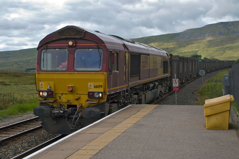 66199, 12.40 Newbiggin-Arpley-West Burton (4B13), Ribblehead station 
 With some welcome sunshine just catching the fells in the background but not the passing train 66199 passes Ribblehead station. It is leading the 4B13 12.40 12.40 Newbiggin to West Burton power station gypsum empties. Ribblehead station closed in May 1970 as the line was being run down as plans for closure took shape but reopened again in 1986 following the saving of the line. Today, the station is leased by the Settle and Carlisle Railway Trust and is put to a variety of uses including holiday accommodation, a museum and a caf. 
 Keywords: 66199 12.40 Newbiggin-Arpley-West Burton gypsum 4B13 Ribblehead station