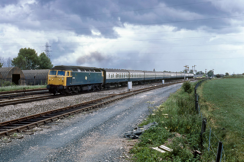 47182, 08.58 Derby-Newquay (1V70), Silk Mill crossing 
 In the 1970/80s a train heading west on a summer Saturday composed of a long rake of Mk.Is was either a relief for a Paignton or a Newquay service. The latter is the case here as 47182 leads the 08.58 Derby to Newquay 1V70 service past Silk Mill crossing just west of Taunton. This view is still possible today but access to the land in the foreground is strictly not allowed due to it being part of the huge Fairwater Yard PW facility. The wartime huts to the left are long gone with the land now occupied by the Bindon Road business park. 
 Keywords: 47182 08.58 Derby-Newquay 1V70 Silk Mill crossing