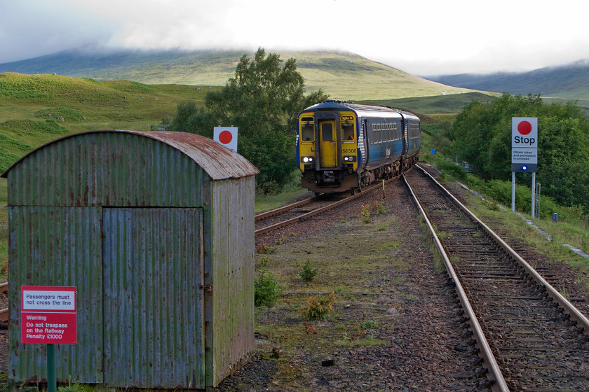 156500, SR 06.03 Mallaig-Glasgow Queen Street (1Y42), Rannoch station 
 With a typical Highland backdrop, 156500 arrives into Rannoch station with the 06.03 Mallaig to Glasgow Queen Street. I deliberately chose to include the wartime style corrugated iron hut off the platform end,. There are a number of these scattered about on this section of line that I suspect contains various maintenance equipment. Obviously, they are of this utilitarian but very practical design to withstand the rigours of the extreme winter weather experienced at locations such as this at just over a thousand feet above sea level. Also, notice the large RETB stop boards indicating a change of section controlled by the signalling Centre at Banavie just west of Fort William. 
 Keywords: 156500 06.03 Mallaig-Glasgow Queen Street 1Y42 Rannoch station ScotRail Sprinter