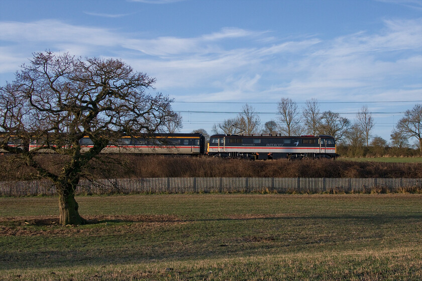 87002, outward leg of The Clansman, 06.58 London Euston-Inverness (1Z42, 4E), Milton crossing 
 The blue sky and the early sunshine belies the fact that it was a bitterly cold morning making the wait for this train a little uncomfortable being exposed in this field! 87002 'Royal Sovereign' makes a fine sight as it passes between Roade and Blisworth on the 'old' fast line via Weedon leading the outward leg of the two-day Clansman charter from Euston to Inverness running as 1Z42. The AC electric is leading a matching set of stock wearing a reproduction version of the very smart InterCity Executive livery that probably marked the pinnacle of BR's ever-changing branding programme. 
 Keywords: 87002 The Clansman 06.58 London Euston-Inverness 1Z42 Milton crossing InterCity Swallow Royal Sovereign