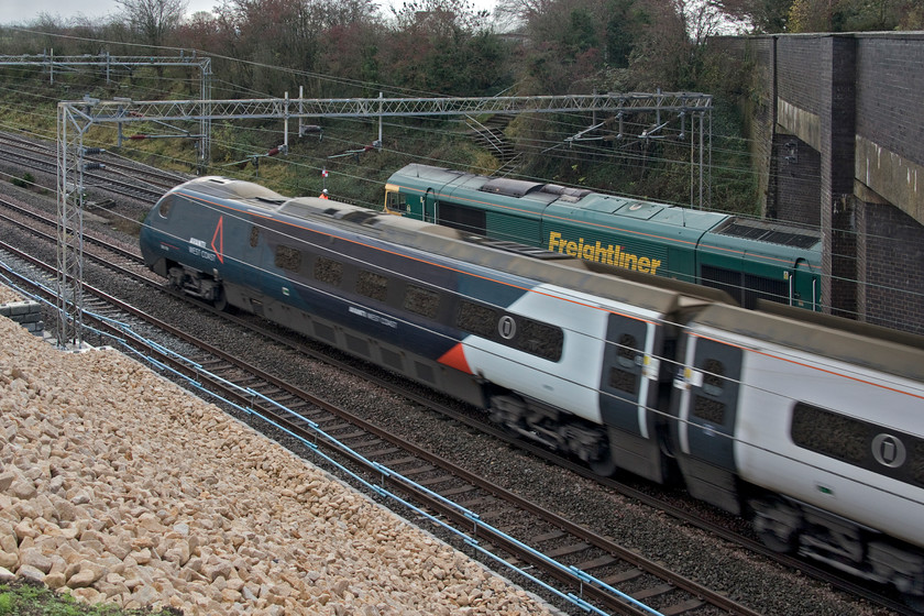 Class 390, VT 09.16 London Euston-Liverpool Lime Street (1F11, 5L) & 66553, engineering train after overnight possession at Blisworth, Ashton Road bridge 
 With the possession still in place on the fast lines (the Weedon loop) all trains were being diverted via Northampton. An unidentified Class 390 Pendolino passes Ashton Road bridge near Roade working the 09.16 Euston to Liverpool Lime Street train. Meanwhile, a mobile signalman under the control of the signalling centre holds 66553 leading a long engineering train following a possession of the fast lines over the previous night. Soon, the Class 66 would be released to make its way under red signals to Hanslope Junction where it would pass from the fast to the slow lines and head to Bletchley to run round and layover for an hour or so. The fast lines could then be handed back and normal services could resume. 
 Keywords: Class 390 09.16 London Euston-Liverpool Lime Street 1F11 66553 engineering train possession at Blisworth Ashton Road bridge Avanti West Coast Freightliner