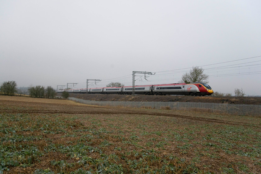 Class 390, VT 06.55. Manchester Piccadilly-London Euston (1A08), Blisworth 
 Recent clearance of the embankments here at Blisworth has enabled photographs from the adjacent field to be taken with just the palisade fencing spoiling the views. A class 390 Pendolino speeds southwards through the winter gloom forming the 06.55 manchester to Euston. My time here at Blisworth is done so it's time to head home for a hearty breakfast! 
 Keywords: 06.55. Manchester Piccadilly-London Euston 1A08 Blisworth