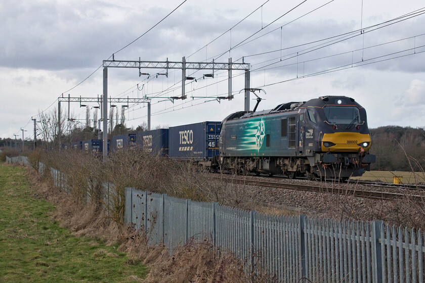 88002, 13.51 DIRFT-Tilbury (4L48, 2E), Wilson's crossing 
 With the sun now firmly hidden by some unexpected cloud blowing in from the northwest 88002 'Prometheus' approaches Northampton at Wilson's crossing with the daily 4L48 Daventry to Tilbury Tesco Express. This service appears to be electrically hauled pretty much every day since the New Year that makes sense given that it runs under the wires for all of its journey. Following the complete clearance of the land to the left of the track a few years ago, it appears that this coming summer may be the last chance to take photographs at this spot of up trains before it takes over unless it is cleared again. 
 Keywords: 88002 13.51 DIRFT-Tilbury 4L48 Wilson's crossing Tesco Express Prometheus