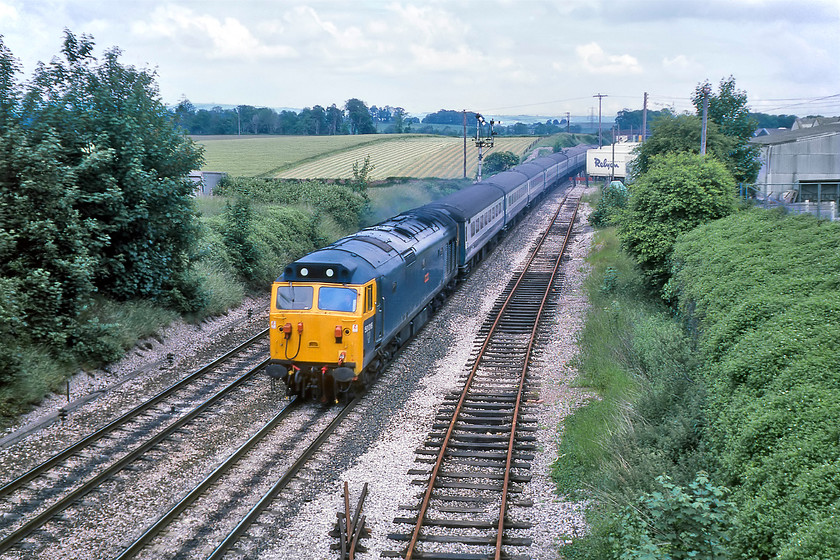 50046, 11.50 London Paddington-Paignton, Wellington 
 50046 'Ajax' is working hard leading the 11.50 Paddington to Paignton summer express as it climbs towards Whiteball summit. It is about to pass under Wellington's B3187 Milverton Road bridge just south of the site of the former station. It is running adjacent to a disused siding leading towards the Reylon factory. I wonder if the siding was used in connection with Reylon's distribution needs in the past; local knowledge anybody? Reylon is still on this site today manufacturing high-quality hand made beds for those people with a little more money to spend! Unfortunately, 50046 is not with us today being cut up by MC Metals in Glasgow during June 1992. 
 Keywords: 50046 11.50 London Paddington-Paignton Wellington Ajax