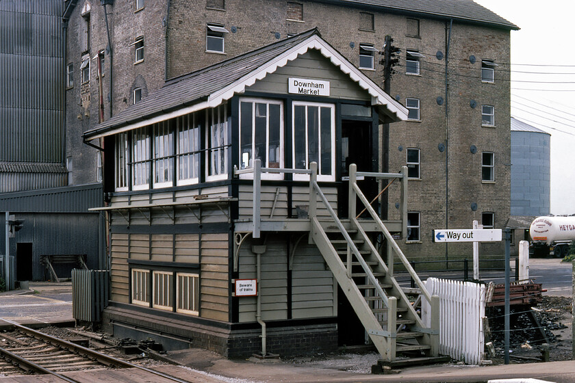 Downham Market signal box (GE, 1881) 
 Downham Markets signal box still stands and, at the time of writing in 2023, is still in use as a crossing box and virtually identical today as per this 1981 photograph. Its a Great Eastern Type 2 box dating from 1881 that stands at the southern end of the down-platform ramp with passengers using a flat crossing to join and leave the platform but today this arrangement has been revised with the exit and entry behind the box. As the box is now listed its future is secure in one form or another when closure eventually comes. The large building that dominates the background is Heygates 1851 built and once steam-powered flour mill. Today, the scene is the same barring a new office building constructed where the Heygates lorry is parked up to the right. 
 Keywords: Downham Market signal box GE Great Eastern Railway