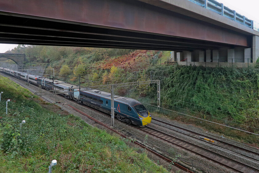 390137, VT. 10.37 Manchester Piccadilly-London Euston (1A19,RT), Roade bypass bridge 
 An extremely wide-angled view of Roade cutting with the new and particularly ugly-looking bridge that carries Roade's new bypass. The comparison with the elegant Victorian brick structure that is my usual haunt is dramatic! 390137 is seen passing away from the camera working the 10.37 Manchester to Euston 1A19 service. Further photographs from this location will soon not be possible as contractors were in the process of fencing off the area beneath the bridge. Notice the line of movement sensors to the left of the image. 
 Keywords: 390137 10.37 Manchester Piccadilly-London Euston 1A19 Roade bypass bridge AWC Avanti West Coast Pendolino