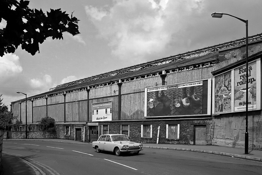 Exterior Bath Green Park station, from Midland Bridge Road 
 The view of Bath Green Park station from Green Park Mews is not as impressive as it is from other angles. The timber cladding is looking a little shabby adorned as it is by period advertising hoardings for Heineken, Butlins holidays and Old Holborn rolling tobacco. There is also a large sign promoting that the station site has been purchased by Sainsbury's and that it is about to be redeveloped. Unfortunately, the DVLA holds no details of the 1969 Mk. 1 Triumph 2000 MFE 555G (originally Lincoln registered) but with its tatty looks I suspect that it did not have many more years on the road! 
 Keywords: Exterior Bath Green Park station from Midland Bridge Road