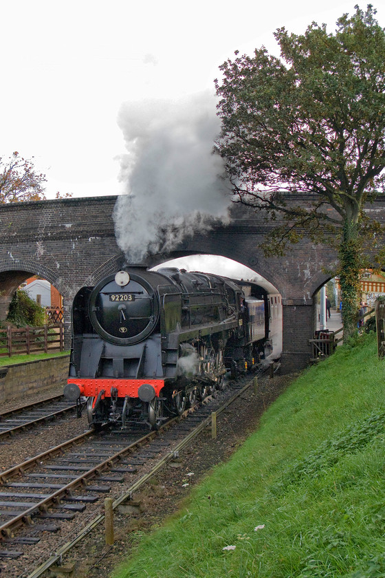 92203, 10.30 Sheringham-Holt, Weybourne 
 With the DMU safely in Weybourne station behind the bridge, 92203 'Black Prince' can get underway and attack Kelling bank working the 10.30 Sheringham to Holt service. In tricky lighting, it gets away from the station making quite a racket as it does. Such a powerful locomotive can make a gentle start away from the station and gather pace relatively easily than smaller and less powerful ones that demand a more determined start from the station.

There is an audio recording of this event on my youtube channel, see.... https://youtu.be/8bmVP9Ytlwc 
 Keywords: 92203 10.30 Sheringham-Holt, Weybourne Black Prince 2-10-0 9F