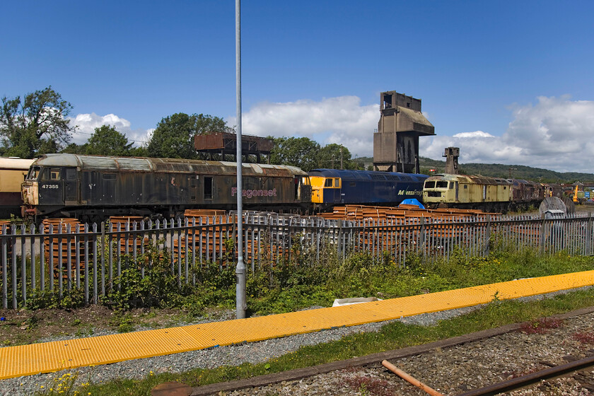 47355, 57005, 47768, 37712, 08418 & 47787, stored, Carnforth WCR 
 A motley collection of first-generation diesels at Carnforth. Wearing its Fragonset livery and branding 47355 leads the lineup with Advenza blue 57005 next. In a rather fetching green livery is 47768 and not 47555 as indicated by the painted number on the cabside. Beyond the green machine is a very tatty 37712, shunter 08418 with 47787 at the far end. Another example of how WCR keep such untidy and poor order. Why have your tattiest and worst assets on full show to the masses like this? To Joe Public, it just looks like a scrapyard which it is, I suppose, in truth!

NB I took a very similar photograph back in 2013, see.... https://www.ontheupfast.com/p/21936chg/30061915097/x3-47500-general-view-carnforth-wcr with just one Class 47 in that view and my predictions about its future came true but it did take seven years before 47500 was cut up! 
 Keywords: 47355 57005 47768 37712 08418 47787 stored Carnforth WCR