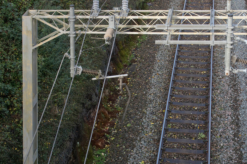 Broken registration arm, Hyde Road bridge 
 A rather sad registration arm hangs limply down having had its contact wire ripped away that can be seen lying in the four foot. The small ash tree that caused this damage is the other side of Roade's Hyde Road bridge that I am standing on looking down on to the track. Ironically, many of the thousands of passengers inconvenienced by this extreme weather event that did this damage, and lots of others for that matter, were delegates travelling to the COP26 climate conference in Glasgow! 
 Keywords: Broken registration arm Hyde Road bridge