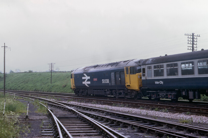 50038, 07.38 Exeter St. David's-London Waterloo (1O06), Whimple goods yard 
 I am not a particular fan of 'going-away shots' but this particular one of 50038 'Formidable' leaving the Devon village of Whimple is interesting. Leading the 1O06 07.38 Exeter St. David's to Waterloo the Class 50 is passing the entrance to Whimple's good yard that still saw occasional use and was largely intact. Notice the telegraph poles carrying the wires that linked the signal boxes that were still in control of this once very important trunk route. By this stage in 1981, its importance was much diminished with long blocks between signal boxes and with it singled for large sections. It was earmarked for closure by BR but remarkably it survived and is very much on the ascendency. 
 Keywords: 50038 07.38 Exeter St. David's-London Waterloo 1O06 Whimple goods yard Formidable