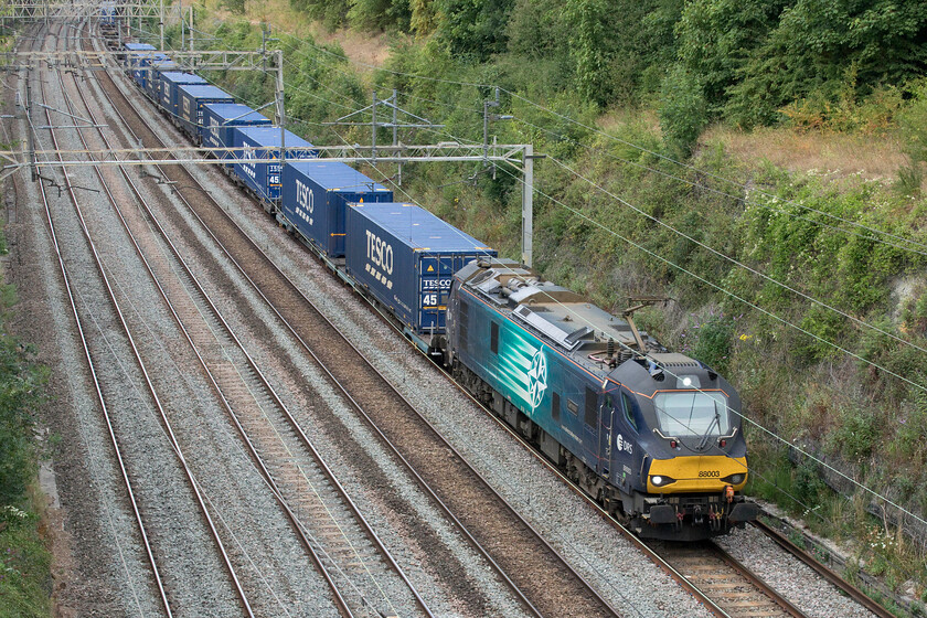 88003, 13.51 DIRFT-Tilbury (4L48, 2E), Hyde Road bridge 
 The weekday 4L48 13.51 Daventry to Tilbury has largely gone over to electric haulage but the weekend services (when they run) are usually in the hands of a DRS Class 66. 88003 'Genesis' leads the service past Roade's Hyde Road bridge making easy work of its train on this particular day. 
 Keywords: 88003 13.51 DIRFT-Tilbury 4L48 Hyde Road bridge DRS Genesis