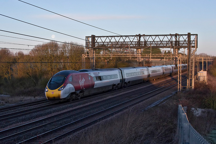 390042, VT 06.43 Manchester Piccadilly-London Euston (1R17, 1L), Roade Hill 
 With the sun just beginning to rise, 390042 passes Roade Hill in Northamptonshire with the 06.43 Manchester to London Euston. Notice the remnants of the decaying super moon from two nights previously that can be seen though the wires. It may not look it, but I can assure you that it was very cold morning, particularly as the warming rays of the rising sun had not quite reached where I was standing yet! 
 Keywords: 390042 06.43 Manchester Piccadilly-London Euston 1R17 Roade Hill