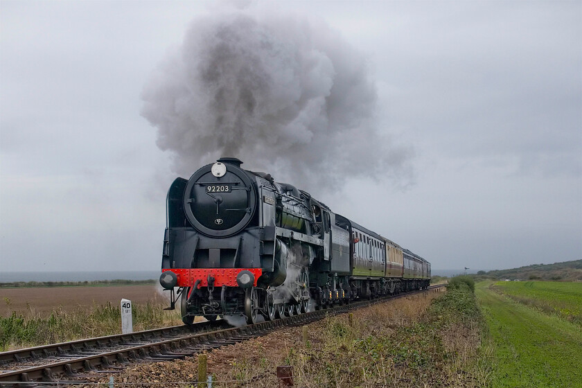 92203, 10.15 Sheringham-Holt, Priory Crossing TG122422-19.10.23 
 Despite being a dull day 92203 'Black Prince' makes a fine sight as it climbs away from the North Sea coast leading the 10.15 Sheringham to Holt service. I have used this spot in the fields just east of Weybourne known as Priory Crossing in the past liking it due to the added interest of the sea in the background and the simple trackside forty-and-a-quarter milepost. The milepost indicates the distance to King's Lyn's M & GN former South Lyn station. 
 Keywords: 92203 10.15 Sheringham-Holt Priory crossing TG122422 Black Prince