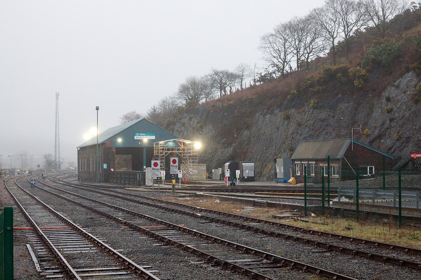 Machynlleth Depot 
 In the gathering gloom of an early evening, Machynlleth stabling point is seen with no units inside. The walls of the structure are all that remain of the GWR steam shed that has been extensively modified for current day use. Note the tall radio mast used for the transmission of the European Rail Traffic Management System (ERTMS) signalling information. 
 Keywords: Machynlleth Depot