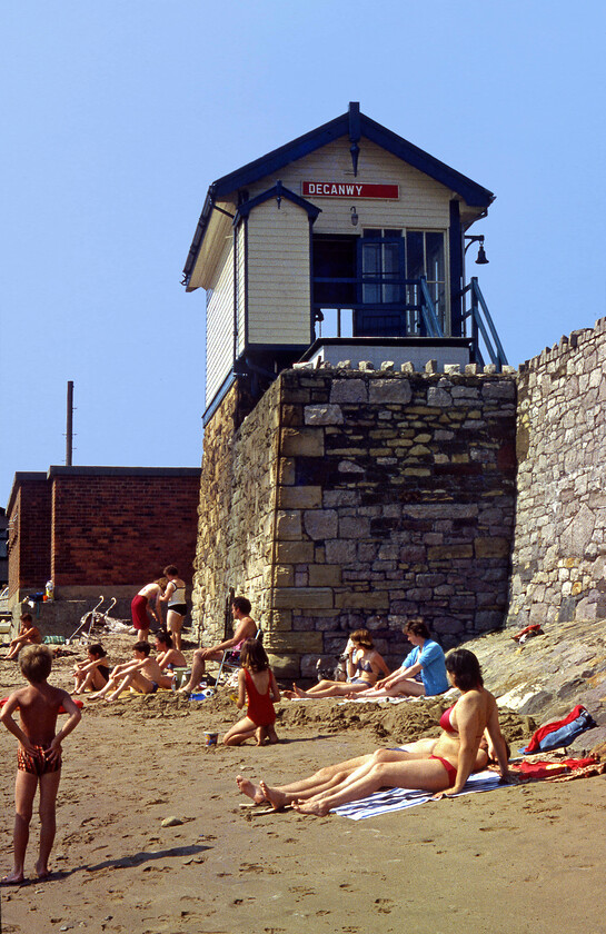 Deganwy signal box (LNW, 1914) 
 The unusually positioned Deganwy signal box is seen from the well-patronised beach on this hot summer Sunday afternoon. The box is a Type 5 L&NWR structure dating from 1914 and was originally named Deganwy Number 2 hence the painted-over section of the wooden post-1935 nameboard. The platform and crossing bell can be seen clearly in this photograph used to warn station staff and pedestrians on the level crossing just beyond the box of the imminent arrival of a train. The visitors to the beach would not have such a pleasant place to sit these days as huge amounts of rock armour have been placed just in front of the sea wall in an effort to retard erosion. 
 Keywords: Deganwy signal box L&NWR