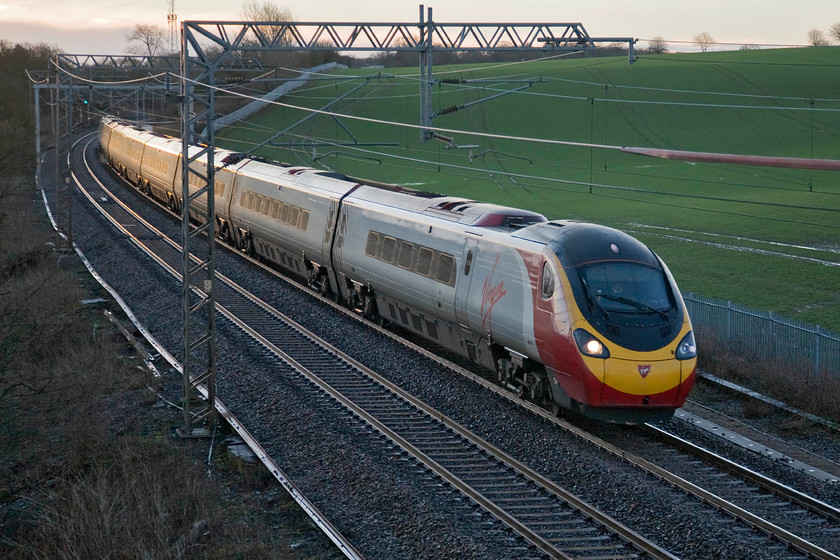 Class 390, VT, unidentified down working, Milton crossing 
 With his disposable cup of coffee placed above the cab controls, the driver brings an unidentified Virgin Pendolino past Milton crossing just north of Roade working a down working from Euston. With the sun just coming over the horizon to the far left the glint lighting is suitably subtle rather than glaring as can often be the case with this type of photograph. 
 Keywords: Class 390 VT unidentified down working, Milton crossing Virgin Trains Pendolino