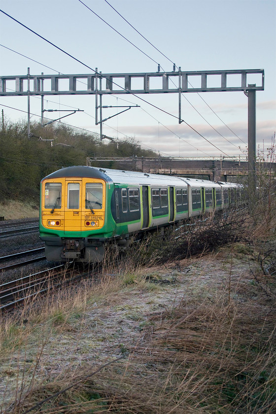 319216, LN 06.50 Northampton-London Euston (2N10, 10L), Ashton Road bridge 
 319216 leads two other units on the 06.50 Northampton to Euston train past Ashton Road bridge just south of the village of Roade. Note the frost on the embankment following a cold night and on what should have been a bright and sunny morning. However, a stubborn area of cloud behind me prevented the sun from illuminating the scene and warming things up! This particular train is a commuter working that is usually very busy. However, due to the Coronavirus outbreak, a quick glance through the windows as it passed revealed that it was almost empty! 
 Keywords: 319216 06.50 Northampton-London Euston 2N10 Ashton Road bridge London Northwestern commuter train