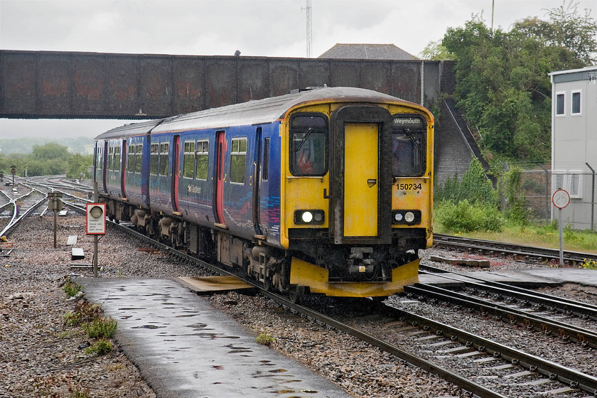 150234, GW 10.42 Gloucester-Weymouth (2O89), Westbury station 
 In the pouring rain, 150234 arrives at Westbury station working the 10.42 Gloucester to Weymouth service. Up until its demolition during the Easter of 1984, Westbury North signal box was located to the left of the unit where the tarmac is. The roof of its replacement, the Westbury PSB, can just be seen above the railway bridge parapet. 
 Keywords: 150234 10.42 Gloucester-Weymouth 2O89 Westbury station First Great Western two car sprinter