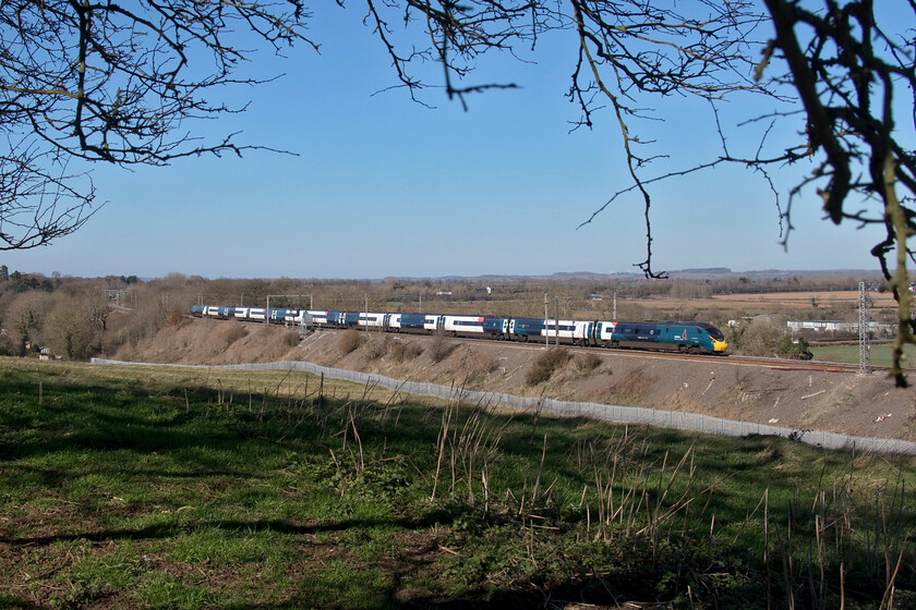 Class 390, VT 10.20 Manchester Piccadilly-London Euston (1A11, 4L), Blisworth 
 Framed by a wizened and windblown tree an unidentified Pendolino heads south past Blisworth working the 1A11 10.20 Manchester to Euston service. The intense blue sky and lush grass contrast with the otherwise pretty colourless end of winter landscape. 
 Keywords: Class 390 10.20 Manchester Piccadilly-London Euston 1A11 Blisworth Avanti West Coast Pendolino