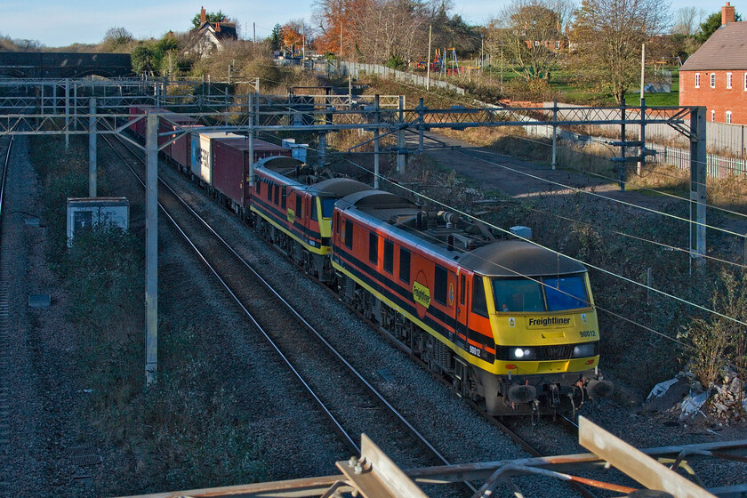 90012 & 90009, 10.20 Trafford Park-Felixstowe North (4L67, 26L), site of Roade station 
 A smart pair of Matching Freightliner locomotives in the form of 90012 and 90009 pass the site of Roade's former station leading the 4L67 10.20 Trafford Park to Felixstowe service. With the low summer sun, today is exactly a month away from the shortest day after all, the scene is struggling for sunshine. Compare this to a photograph taken at the same time of day but in the summer, see... https://www.ontheupfast.com/p/21936chg/29133977204/x323210-10-49-soho-tmd-wolverton 
 Keywords: 90012 90009 10.20 Trafford Park-Felixstowe North 4L67 site of Roade station Freightliner