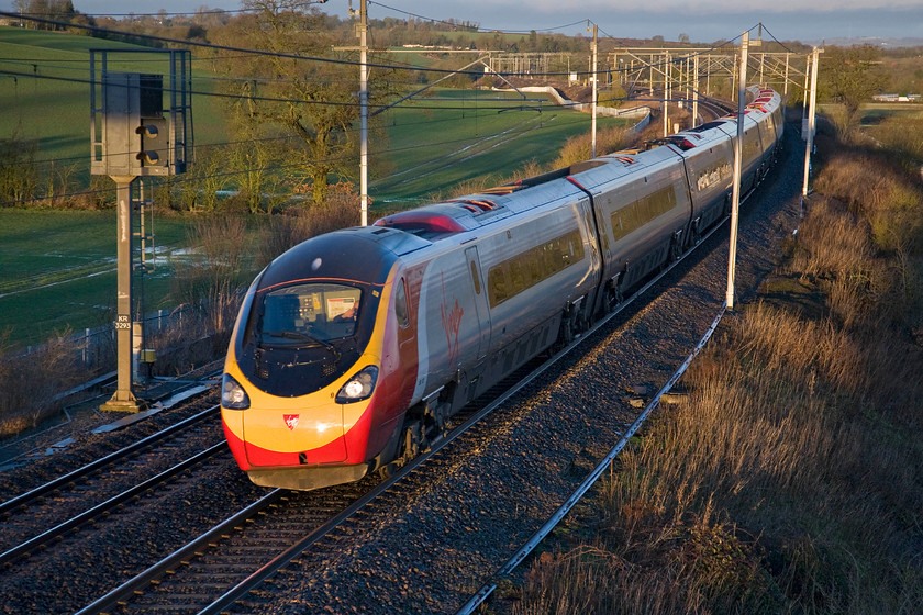 Class 390, VT, unidentified up working, Milton crossing 
 In dramatic early morning lighting, a Virgin Pendolino speeds southwards with an up service for Euston. I am not sure if the driver is shielding his eyes from the rising sun or taking a drink as it sweeps around the curve at Milton crossing on the approach to the north end of Roade cutting. 
 Keywords: Class 390 VT unidentified up working, Milton crossing Virgin West Coast Pendolino