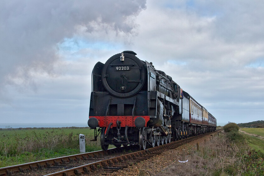 92203, 10.00 Sheringham-Holt, Priory crossing TG122422 
 9F 92203 'Black Prince' hauls the first service train of the day, the 10.00 Sheringham to Holt, past Priory crossing on the approach to Weybourne on Wednesday 19th October 2022. It is quite a climb on this section of the route as the line heads inland away from the sea, seen in the background, but this is no challenge for the might of a 9F! However, later in the morning, something went wrong with 92203 and it was taken out of service with D5631 taking over. Notice the milepost to the left of the locomotive showing forty and a half miles from South Lyn Junction (King's Lyn) via Fakenham on the M&GN route. 
 Keywords: 92203 10.00 Sheringham-Holt, Priory crossing TG122422 Black prince British Railways Standard Class 9F