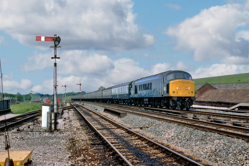 45054, 10.40 Manchester Piccadilly-Paignton (1V85), Tiverton Junction station 
 At last, the sun has come out again on this changeable summer Saturday! 45054 storms through Tiverton Junction leading the 1V85 10.40 Manchester to Paignton train composed of a long rake of Mk.I stock. Notice the unusual appearance of the station loop starter signal in the foreground having lost its finial. Also notice that I have ventured off the platform end and on to the timber barrow crossing to take this picture. This would have been in view of the signalman in the box on the station at the western end of the up platform; how things have changed! 
 Keywords: 45054 10.40 Manchester Piccadilly-Paignton 1V85 Tiverton Junction station